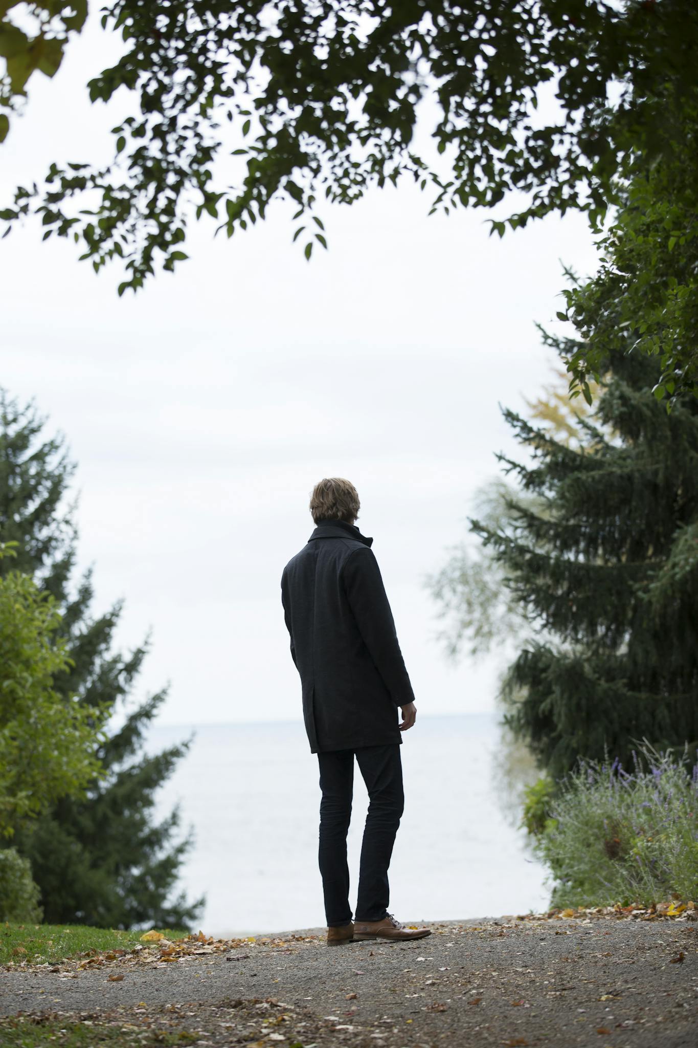 Man in a coat standing on path, surrounded by trees, overlooking a serene lake on a cloudy day.