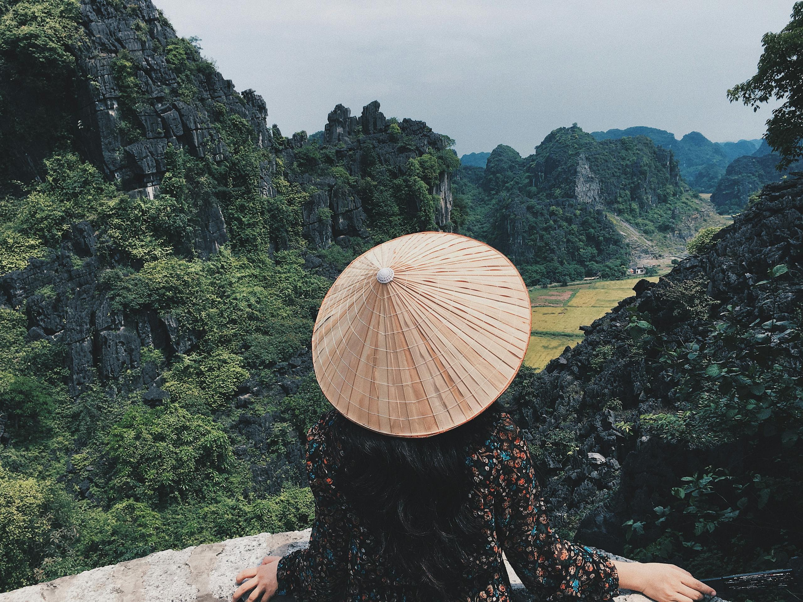 Woman in traditional hat overlooks stunning limestone cliffs in Ninh Binh, Vietnam.